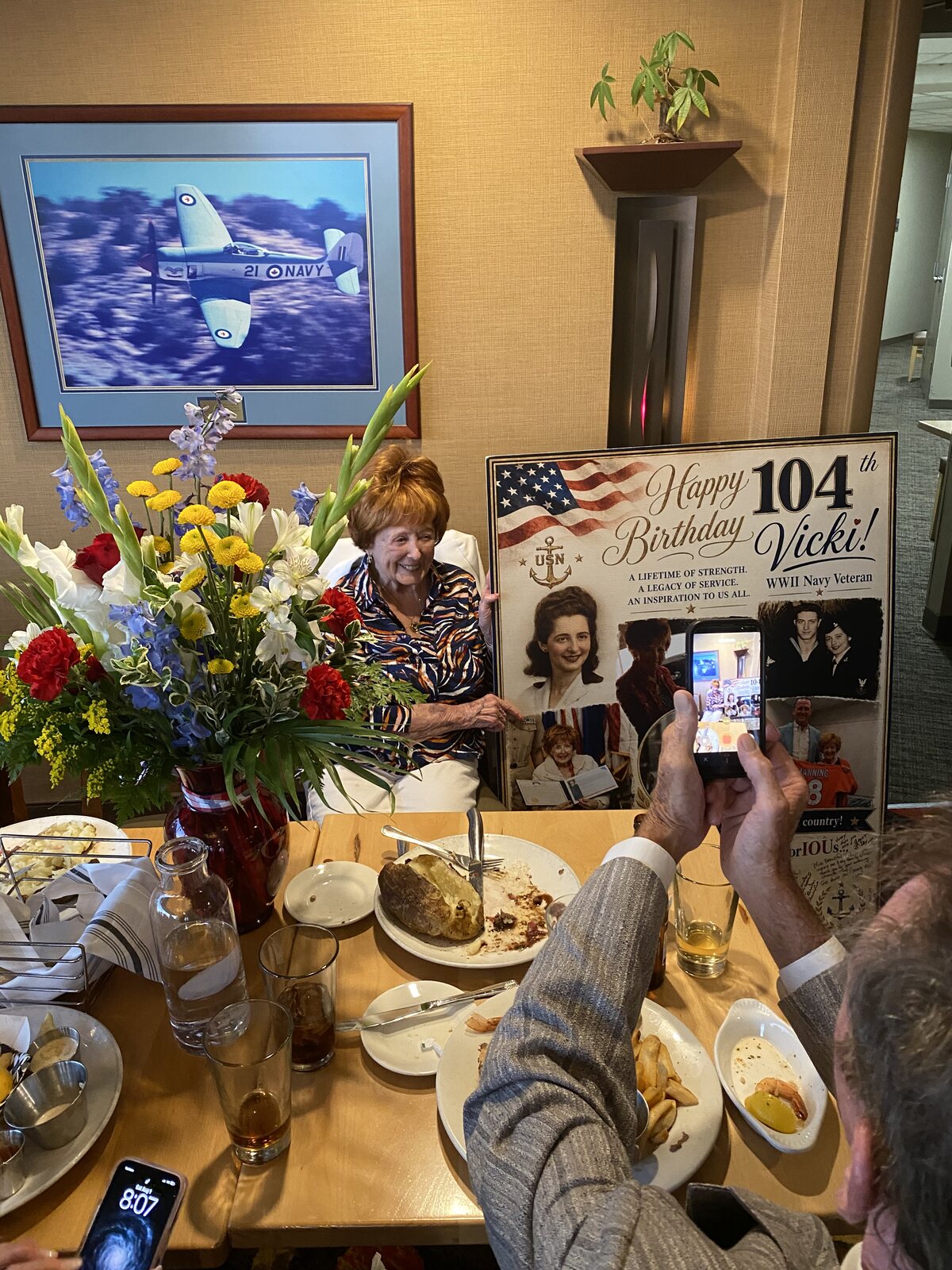 Vicki smiling beside her giant 104th birthday card at the Perfect Landing restaurant, next to a bouquet of flowers