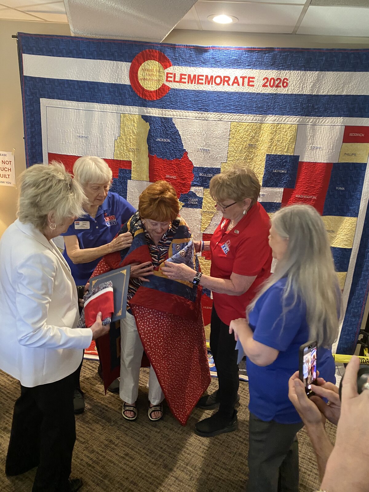 Vicki receiving her Quilts of Valor quilt in front of the Celememorate 2026 Colorado counties quilt